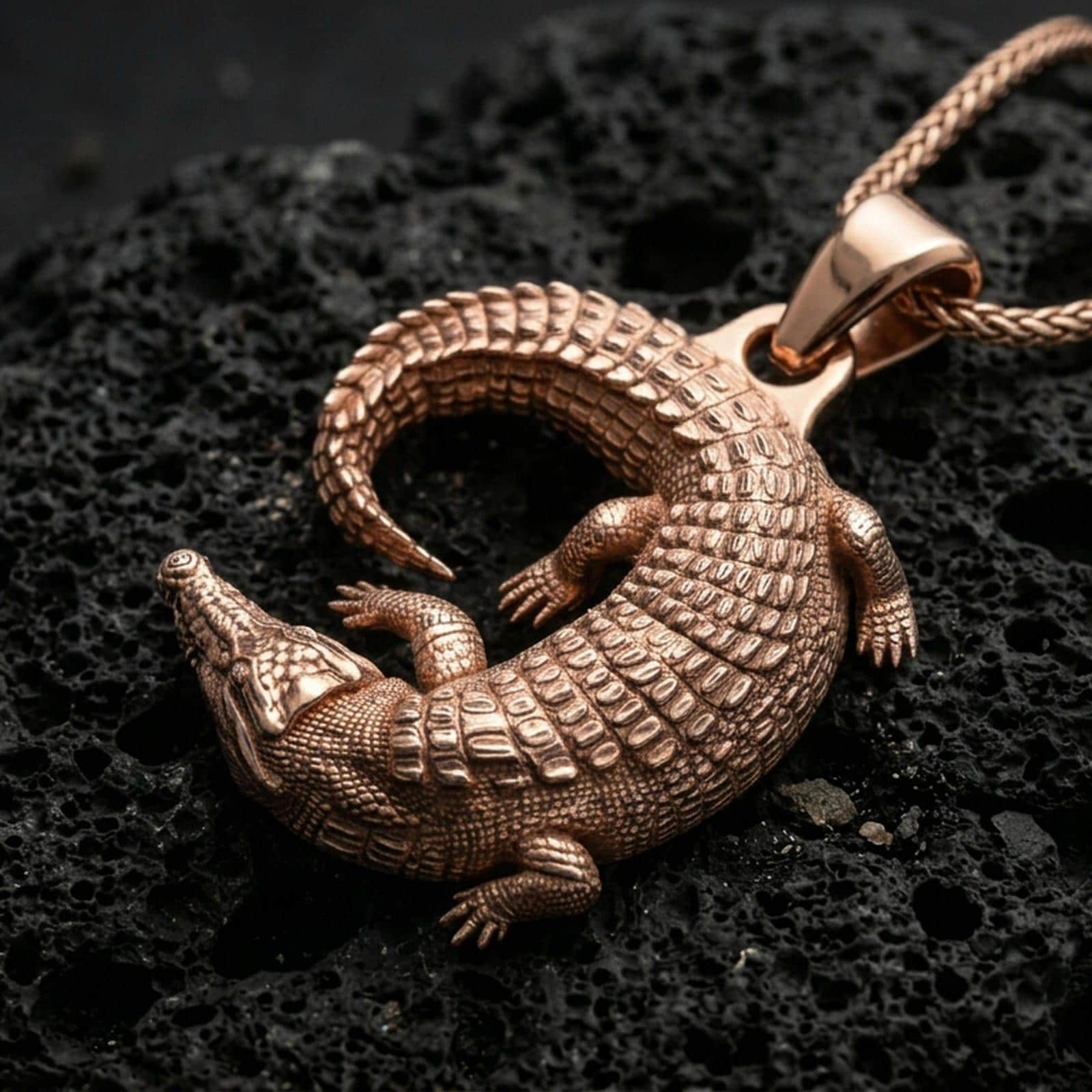 Rose gold plated alligator pendant with realistic reptile relief, textured scale pattern, and dramatic curved silhouette resting on volcanic rock in an elegant close-up product presentation