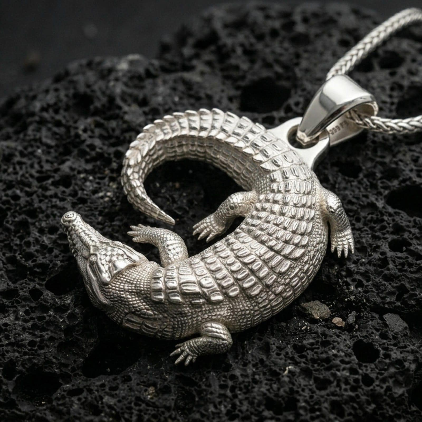 Polished silver alligator necklace with textured scaled body, sculpted reptile form, and lifelike surface detail placed on textured volcanic rock in a premium studio product shot