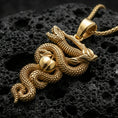 Load image into Gallery viewer, Gold plated serpent pendant with coiled snake body, realistic scale relief, apple motif, and symbolic branch detail showcased on black volcanic rock in a refined macro jewelry image
