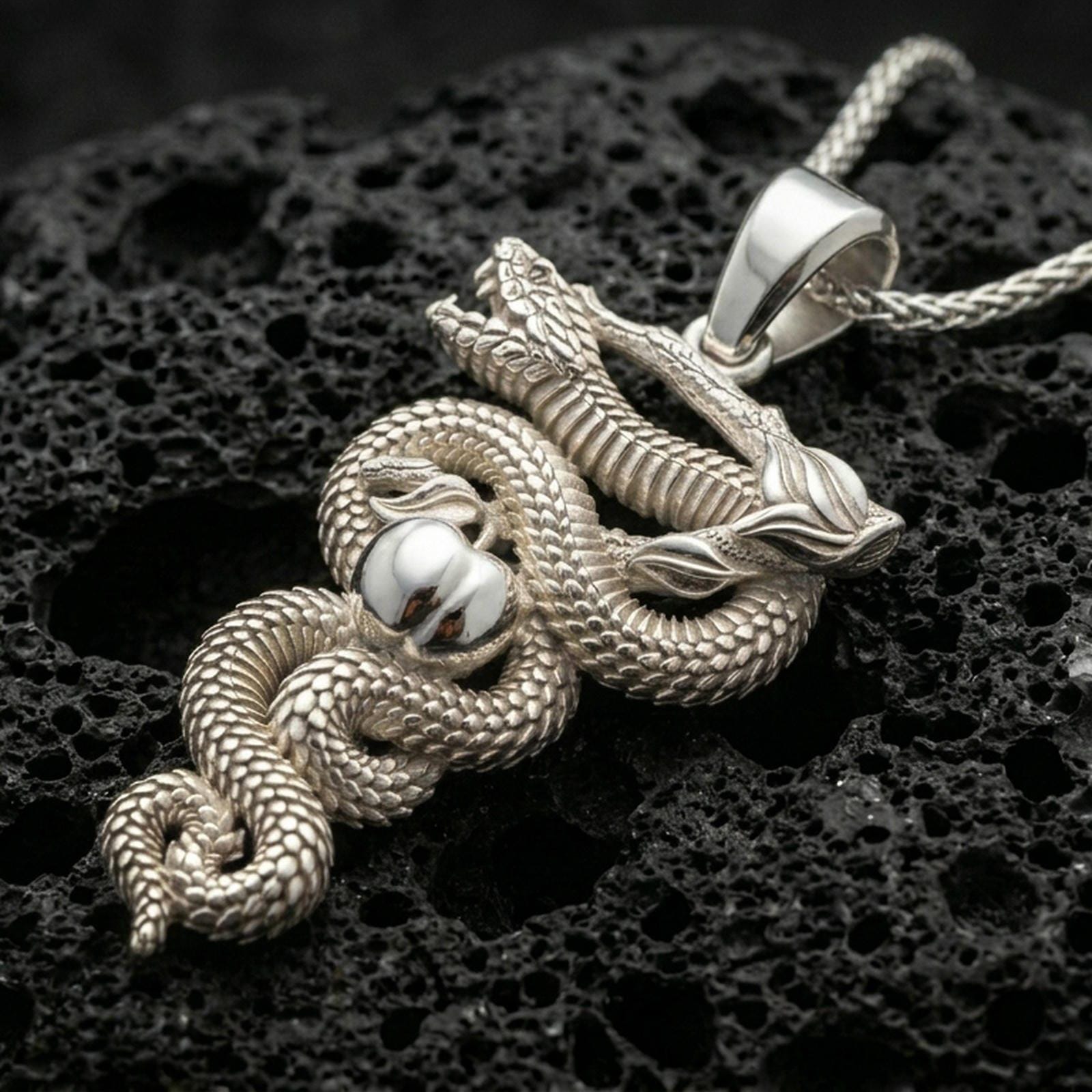 Polished silver snake necklace with detailed scale texture, wrapped serpent form, apple accent, and sculpted branch design placed on textured volcanic rock in a premium studio product shot