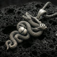 Load image into Gallery viewer, Oxidized serpent pendant with coiled snake, apple detail, and branch motif displayed on dark volcanic rock in a close-up luxury jewelry composition
