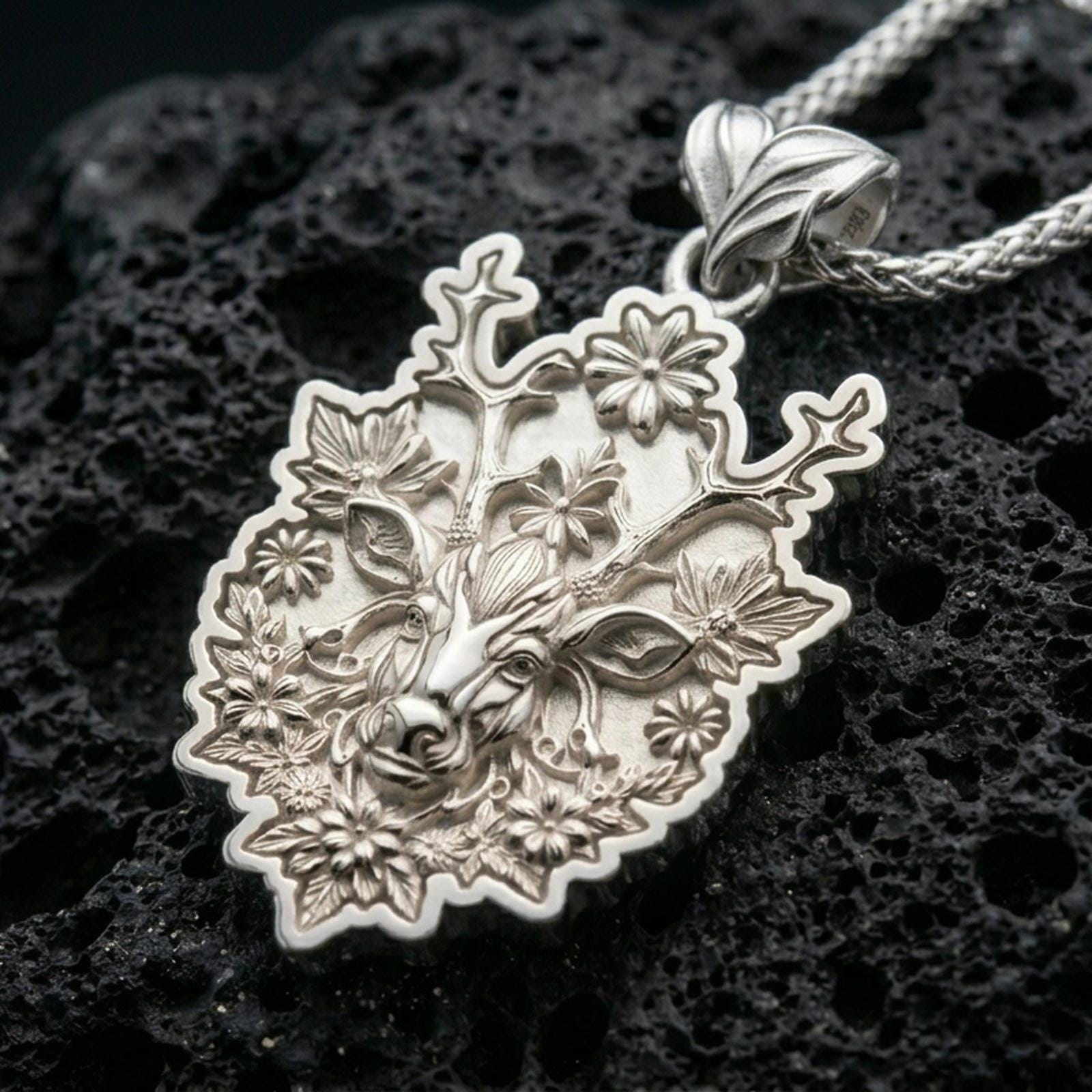 Polished silver stag necklace with blooming flower details, branched antlers, and sculpted woodland animal relief placed on textured volcanic rock in a premium studio product shot