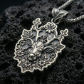 Load image into Gallery viewer, Oxidized deer pendant with floral antlers, detailed stag face, and nature-inspired silhouette displayed on dark volcanic rock in a close-up luxury jewelry composition
