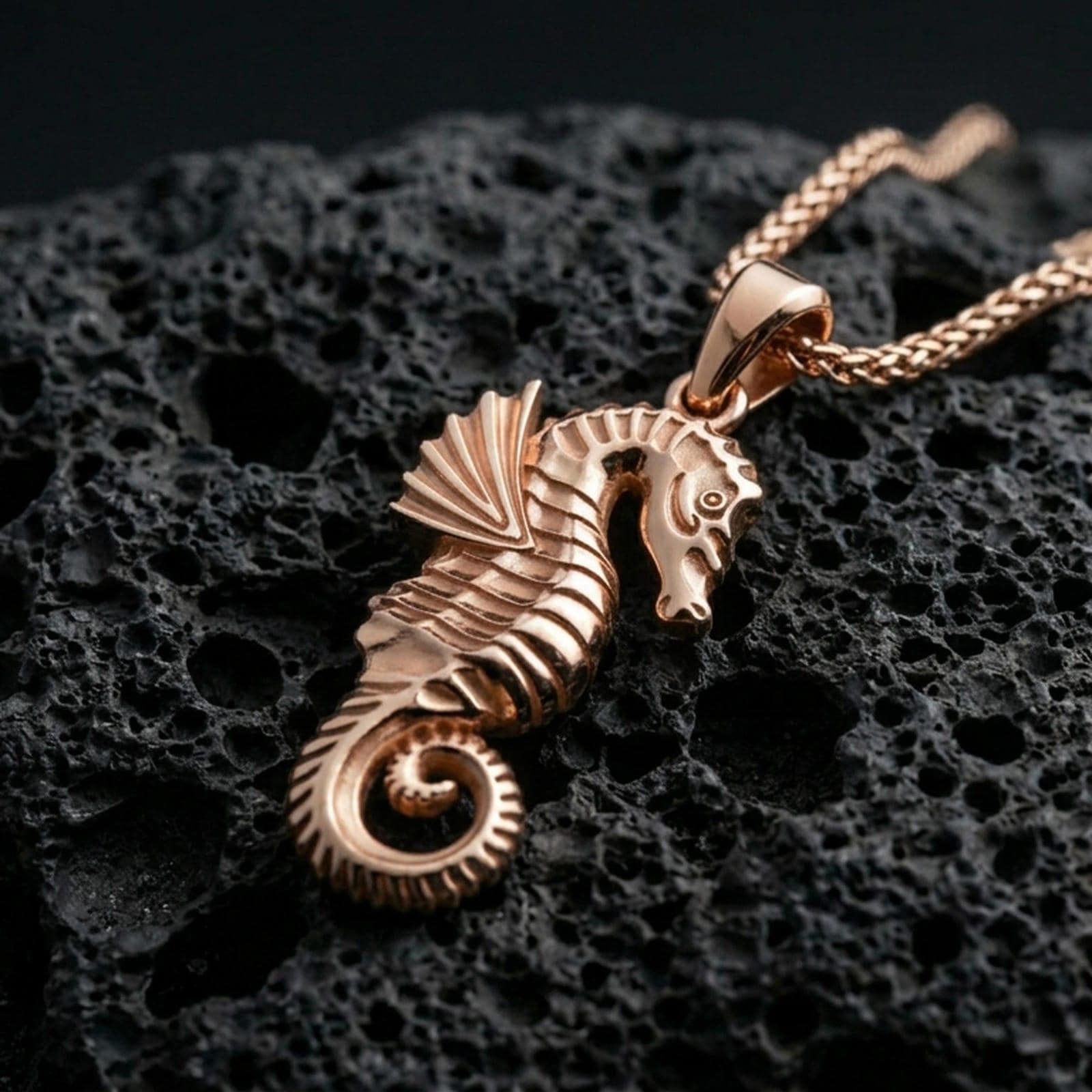 Rose gold plated seahorse pendant with detailed body relief, curled tail, and graceful nautical form resting on volcanic rock in an elegant close-up product presentation
