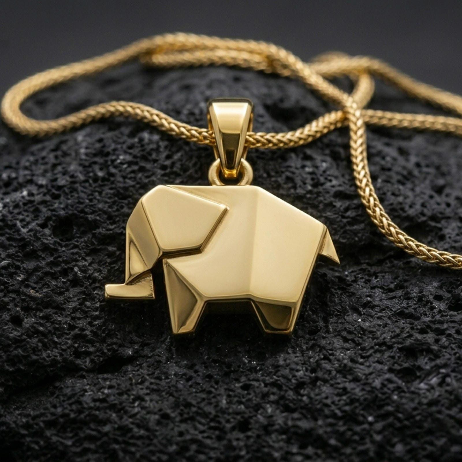 Gold plated origami elephant pendant with geometric folded detailing, minimalist animal shape, and contemporary sculptural form showcased on black volcanic rock in a refined macro jewelry image