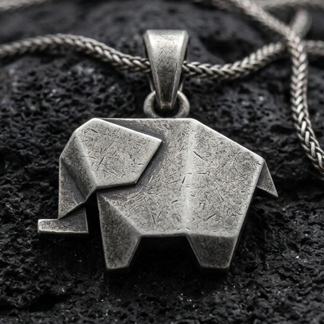Oxidized origami elephant pendant with geometric folded form and minimalist animal silhouette displayed on dark volcanic rock in a close-up luxury jewelry composition