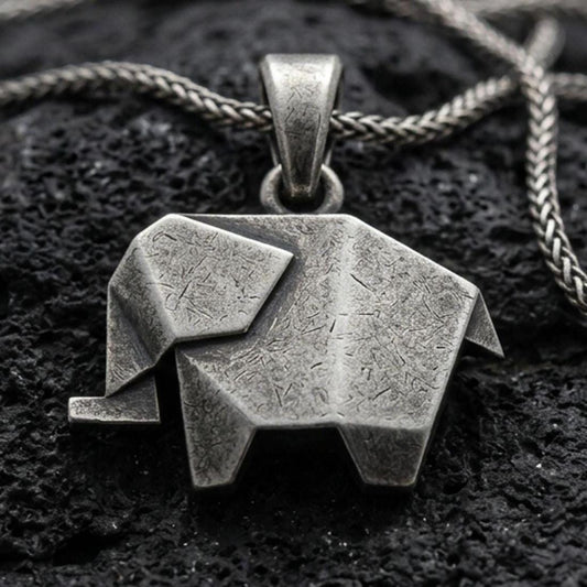 Oxidized origami elephant pendant with geometric folded form and minimalist animal silhouette displayed on dark volcanic rock in a close-up luxury jewelry composition