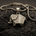 Load image into Gallery viewer, Bronze origami elephant pendant with geometric folded form and minimalist animal silhouette displayed on dark volcanic rock in a close-up luxury jewelry composition
