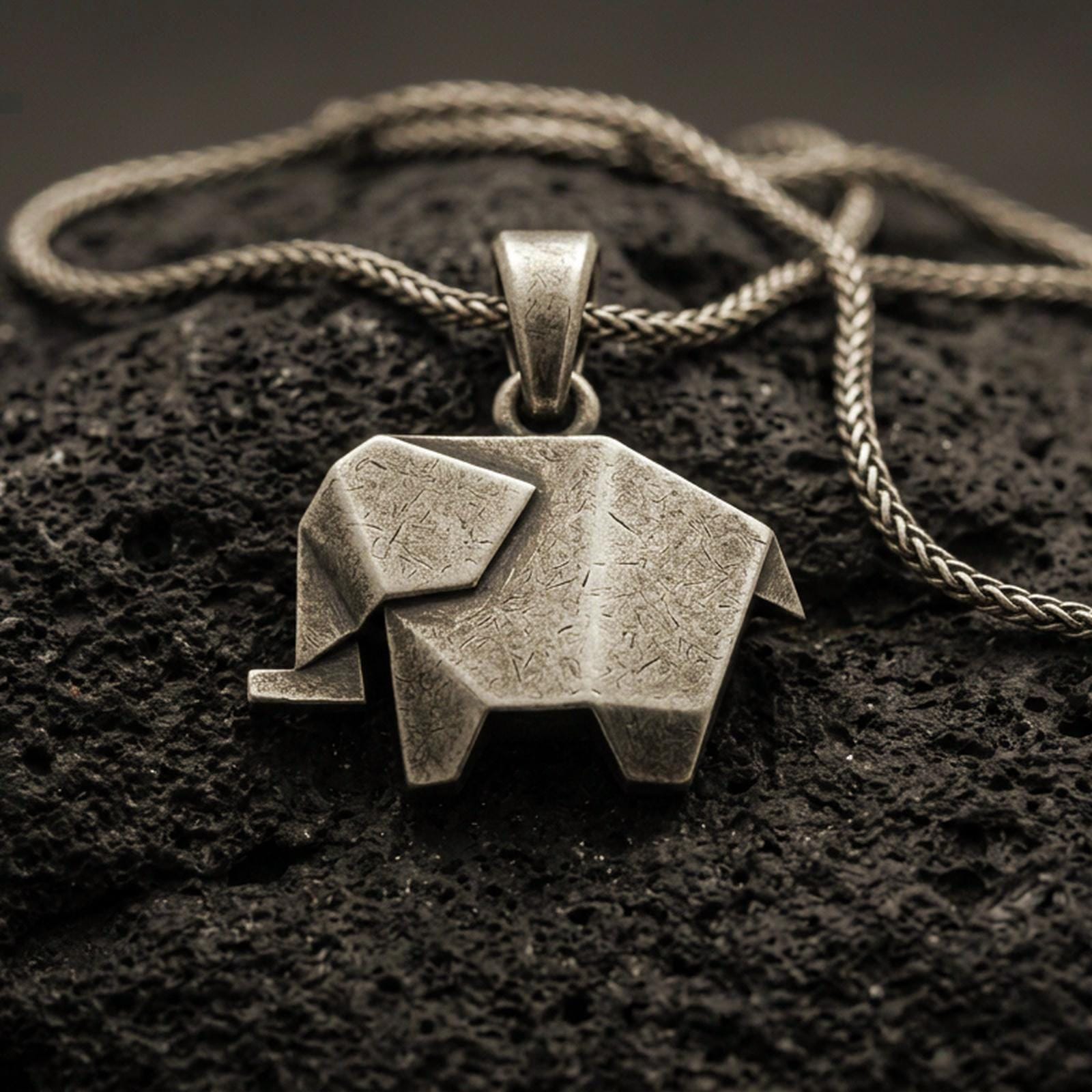 Bronze origami elephant pendant with geometric folded form and minimalist animal silhouette displayed on dark volcanic rock in a close-up luxury jewelry composition