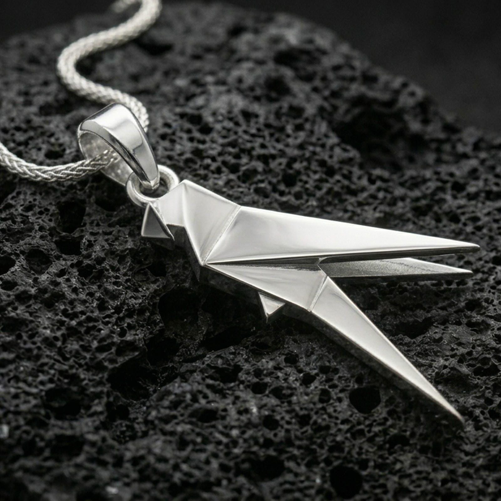 Polished silver parrot necklace with minimalist origami-inspired structure, faceted folded lines, and elongated bird shape placed on textured volcanic rock in a premium studio product shot