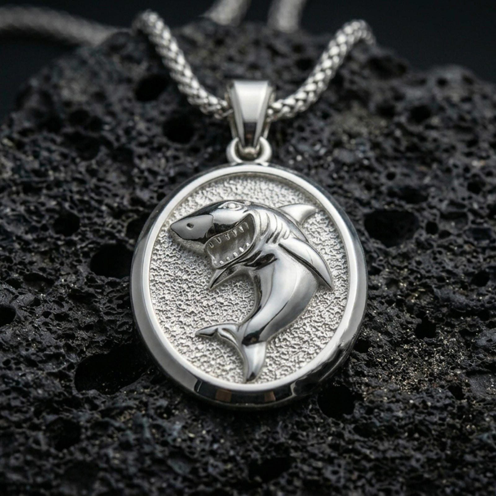Polished silver shark necklace with detailed predatory shark design, oval pendant silhouette, and textured marine-style backdrop placed on textured volcanic rock in a premium studio product shot
