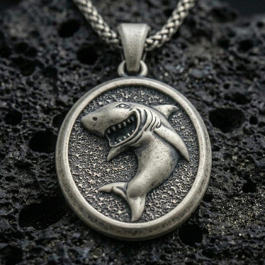 Oxidized shark pendant with fierce open-mouth shark relief, oval medallion frame, and textured ocean-inspired background displayed on dark volcanic rock in a close-up luxury jewelry composition