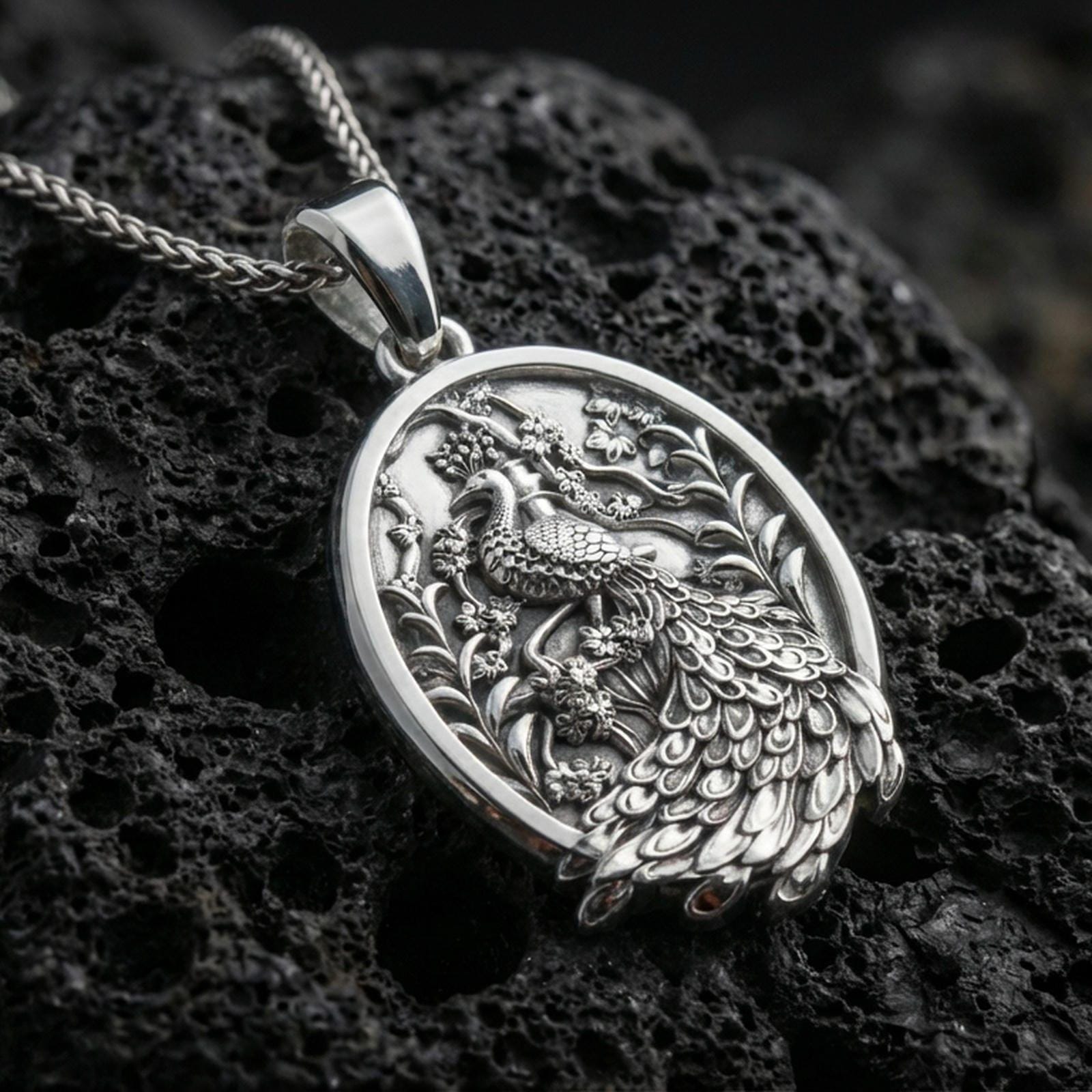 Polished silver peacock necklace with ornate plumage detailing, botanical background elements, and elegant oval frame placed on textured volcanic rock in a premium studio product shot
