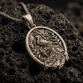 Load image into Gallery viewer, Bronze peacock pendant with detailed feather relief, floral branch accents, and oval medallion silhouette displayed on dark volcanic rock in a close-up luxury jewelry composition
