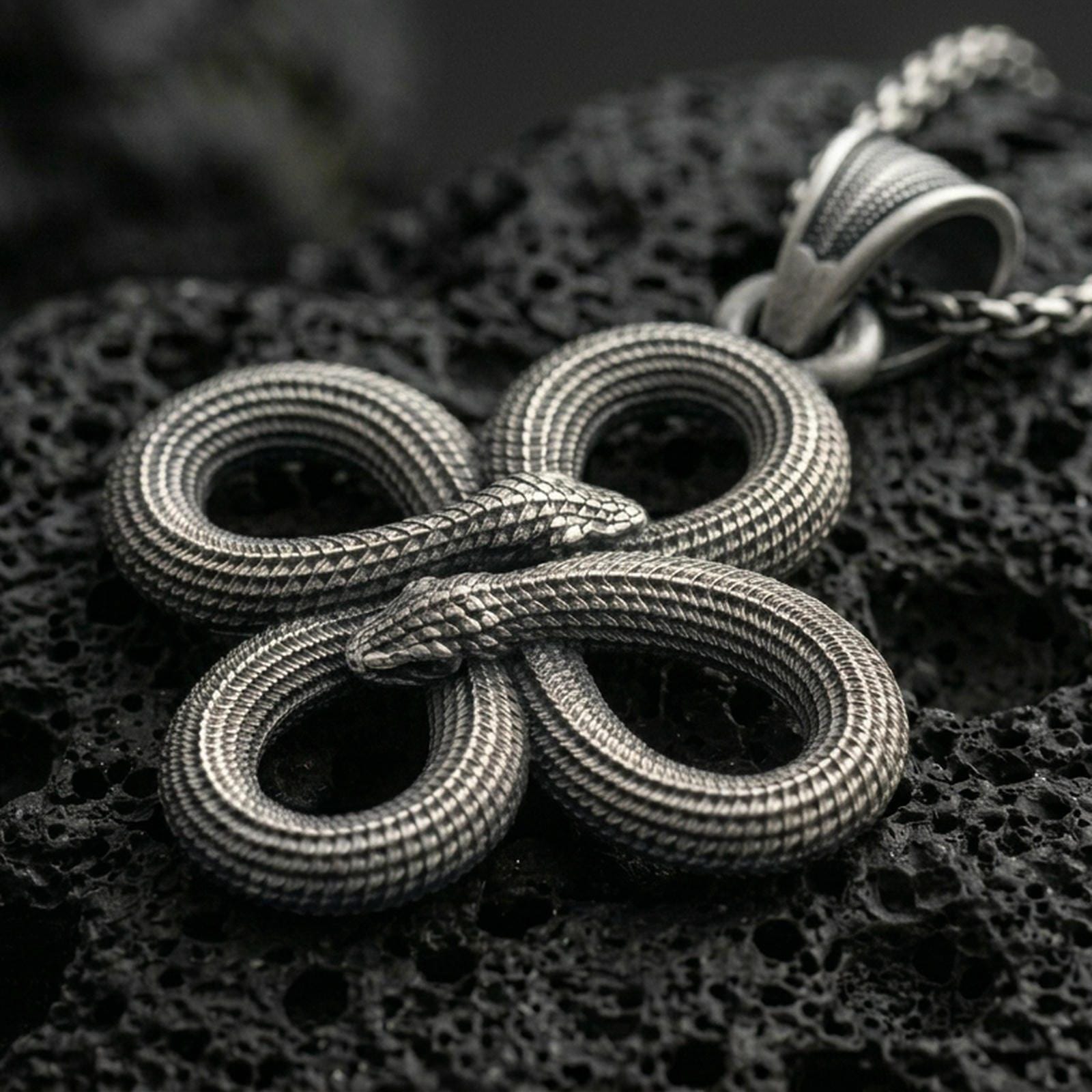 Oxidized snake knot pendant with intertwined serpents, detailed scale texture, and symmetrical looped design displayed on dark volcanic rock in a close-up luxury jewelry composition