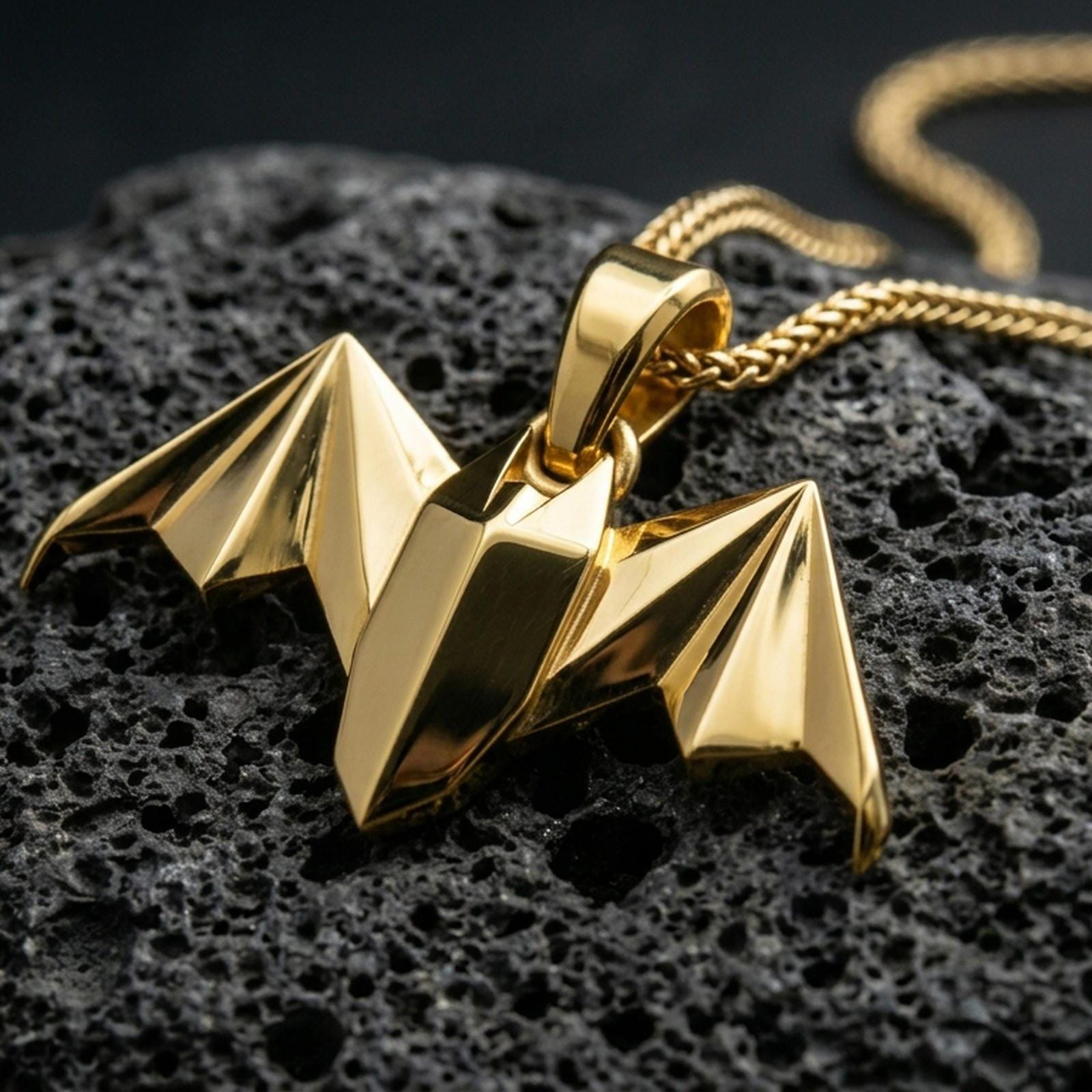 Gold plated origami bat pendant with geometric folded surfaces, pointed wing silhouette, and sleek nocturnal charm showcased on black volcanic rock in a refined macro jewelry image