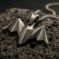 Load image into Gallery viewer, Bronze origami bat pendant with geometric folded wings and sharp nocturnal silhouette displayed on dark volcanic rock in a close-up luxury jewelry composition
