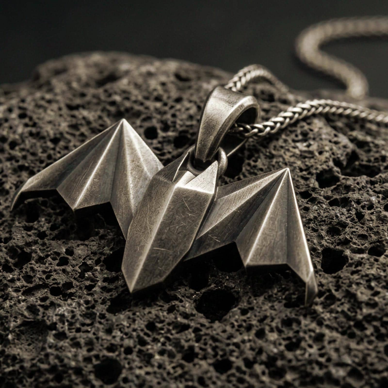 Bronze origami bat pendant with geometric folded wings and sharp nocturnal silhouette displayed on dark volcanic rock in a close-up luxury jewelry composition