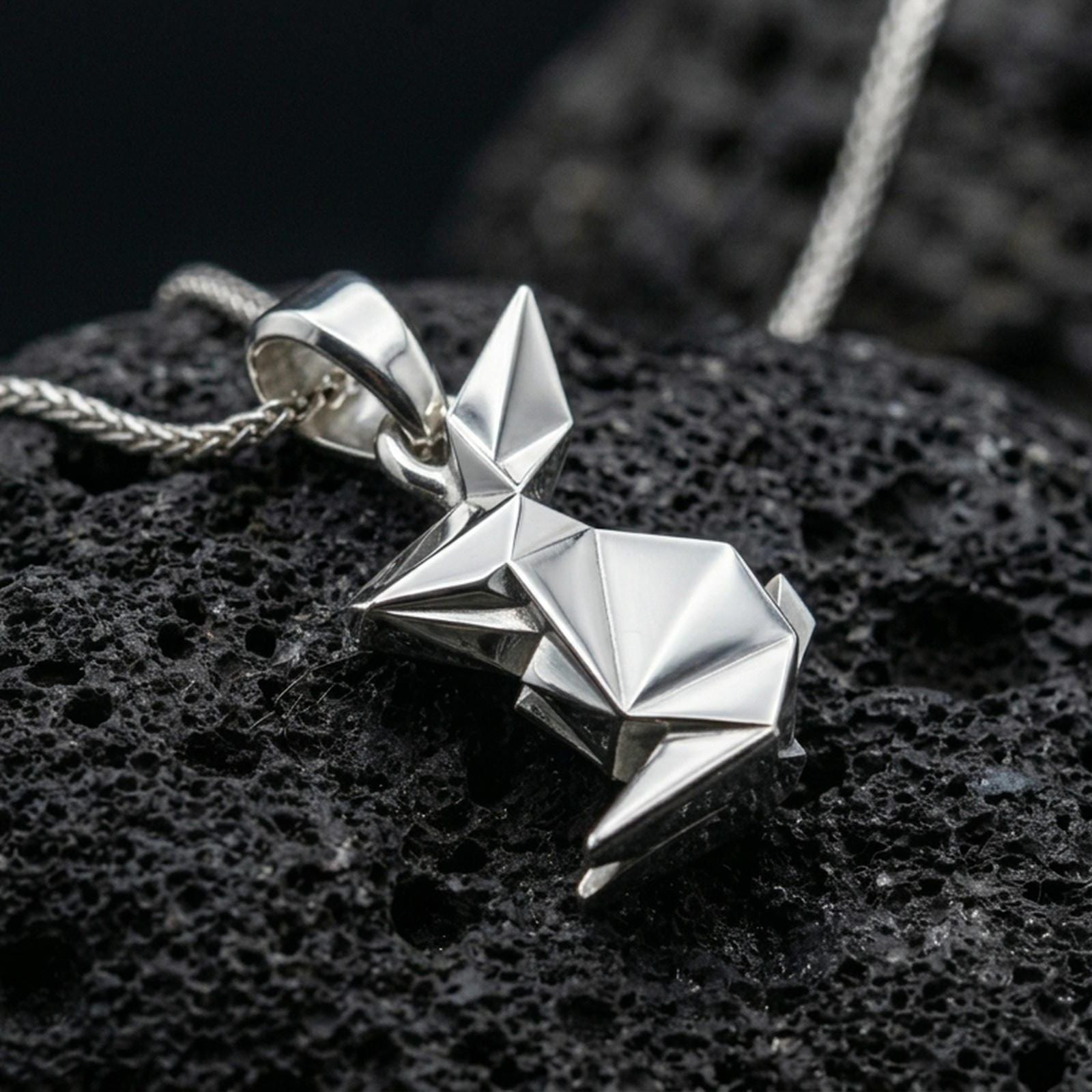 Polished silver rabbit necklace with angular origami-inspired structure, faceted folded surfaces, and modern animal design placed on textured volcanic rock in a premium studio product shot