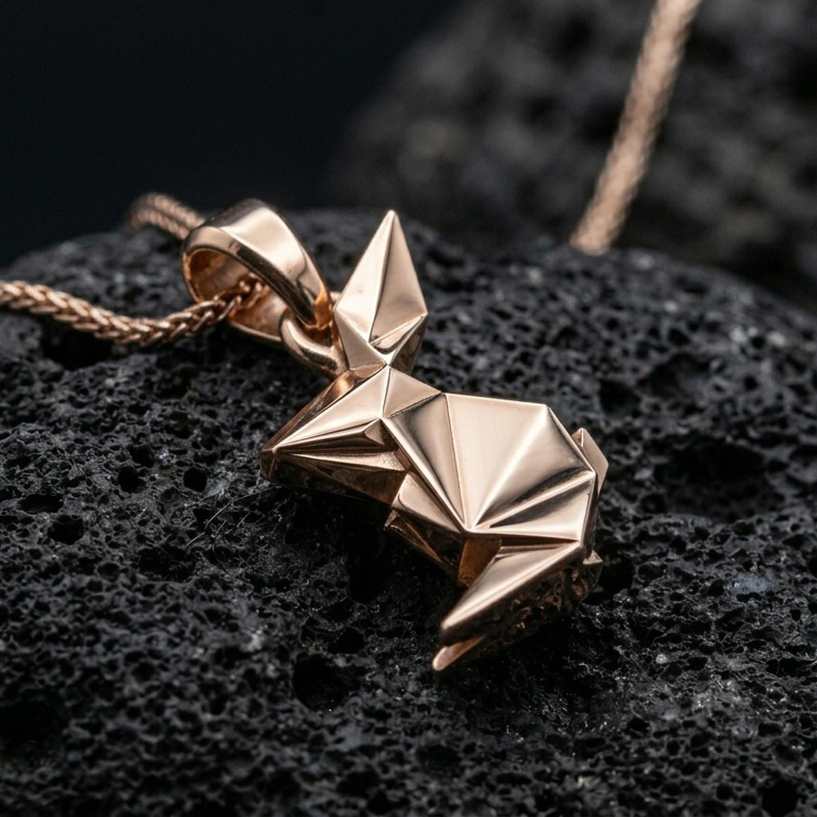 Rose gold plated geometric rabbit pendant with origami-style folds, sleek modern bunny silhouette, and elegant minimalist design resting on volcanic rock in an elegant close-up product presentation