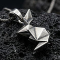 Load image into Gallery viewer, Oxidized origami rabbit pendant with geometric folded form and minimalist bunny silhouette displayed on dark volcanic rock in a close-up luxury jewelry composition
