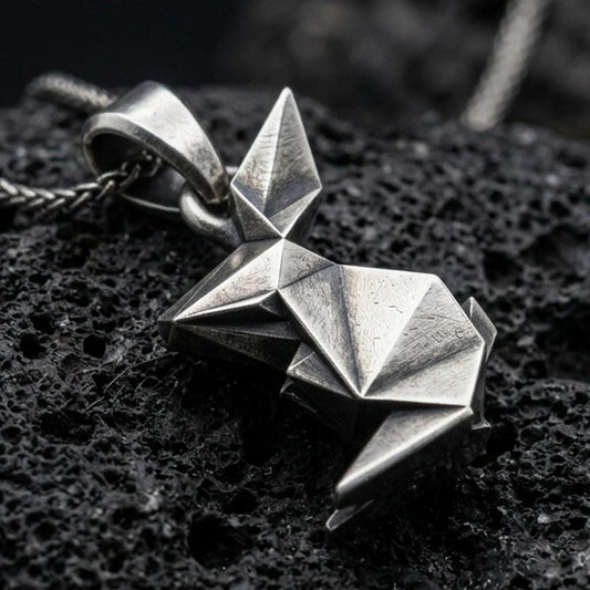 Oxidized origami rabbit pendant with geometric folded form and minimalist bunny silhouette displayed on dark volcanic rock in a close-up luxury jewelry composition