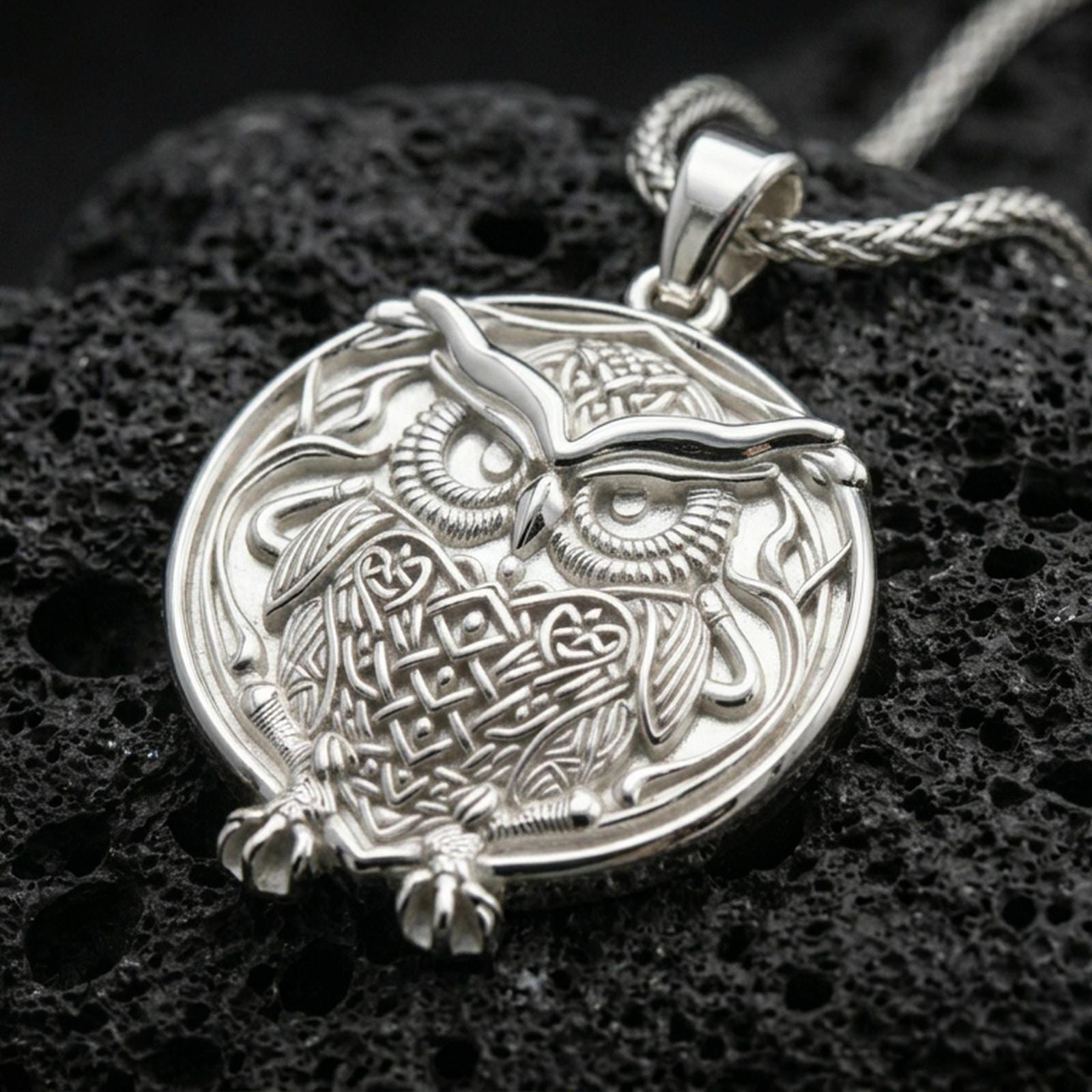 Polished silver owl necklace with intricate feather texture, circular medallion form, and expressive wise bird design placed on textured volcanic rock in a premium studio product shot