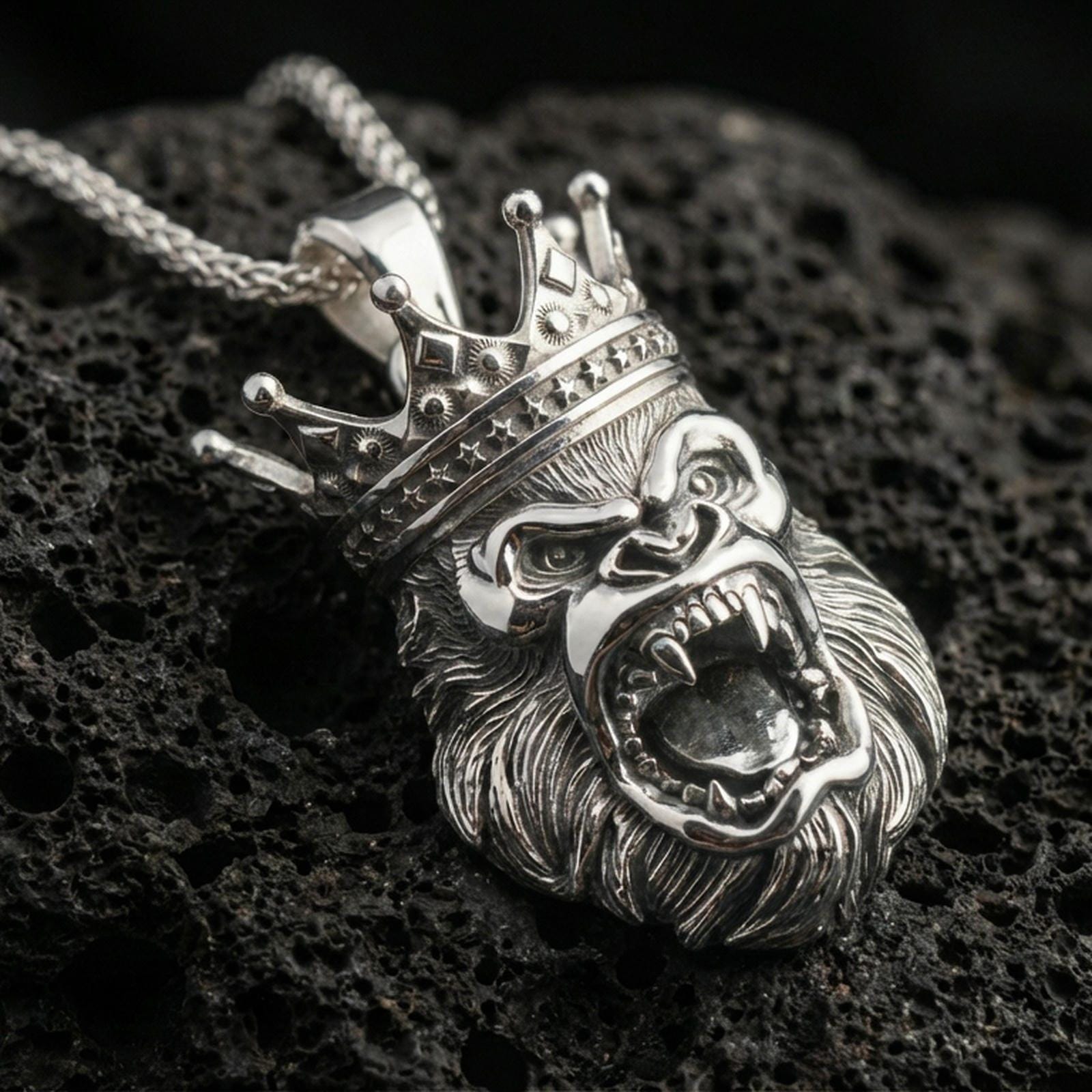 Polished silver gorilla necklace with crowned primate design, fierce open-mouth expression, and sculpted fur detailing placed on textured volcanic rock in a premium studio product shot