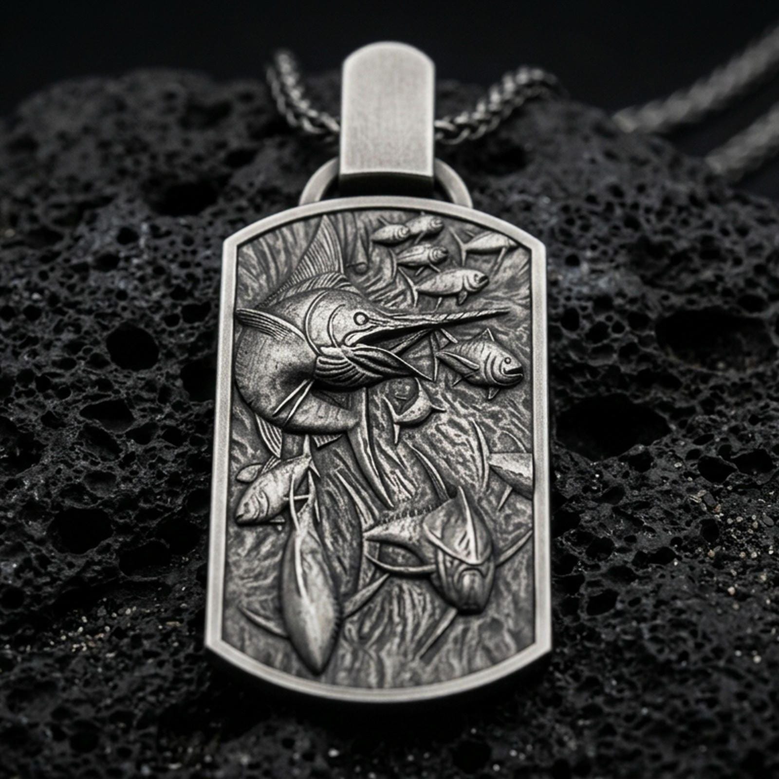 Oxidized swordfish pendant with detailed ocean life scene, swimming fish relief, and rectangular medallion silhouette displayed on dark volcanic rock in a close-up luxury jewelry composition