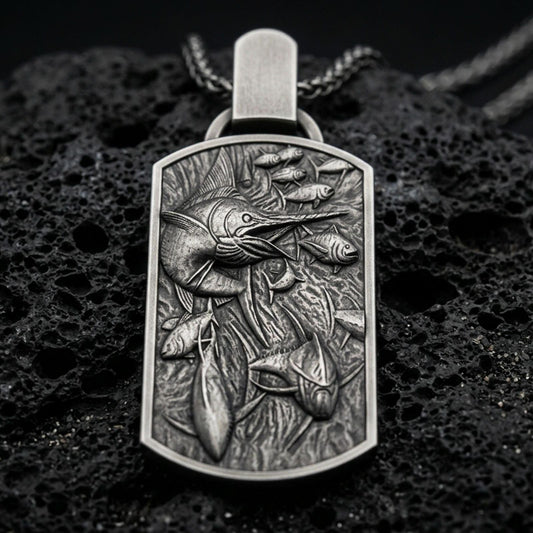 Oxidized swordfish pendant with detailed ocean life scene, swimming fish relief, and rectangular medallion silhouette displayed on dark volcanic rock in a close-up luxury jewelry composition
