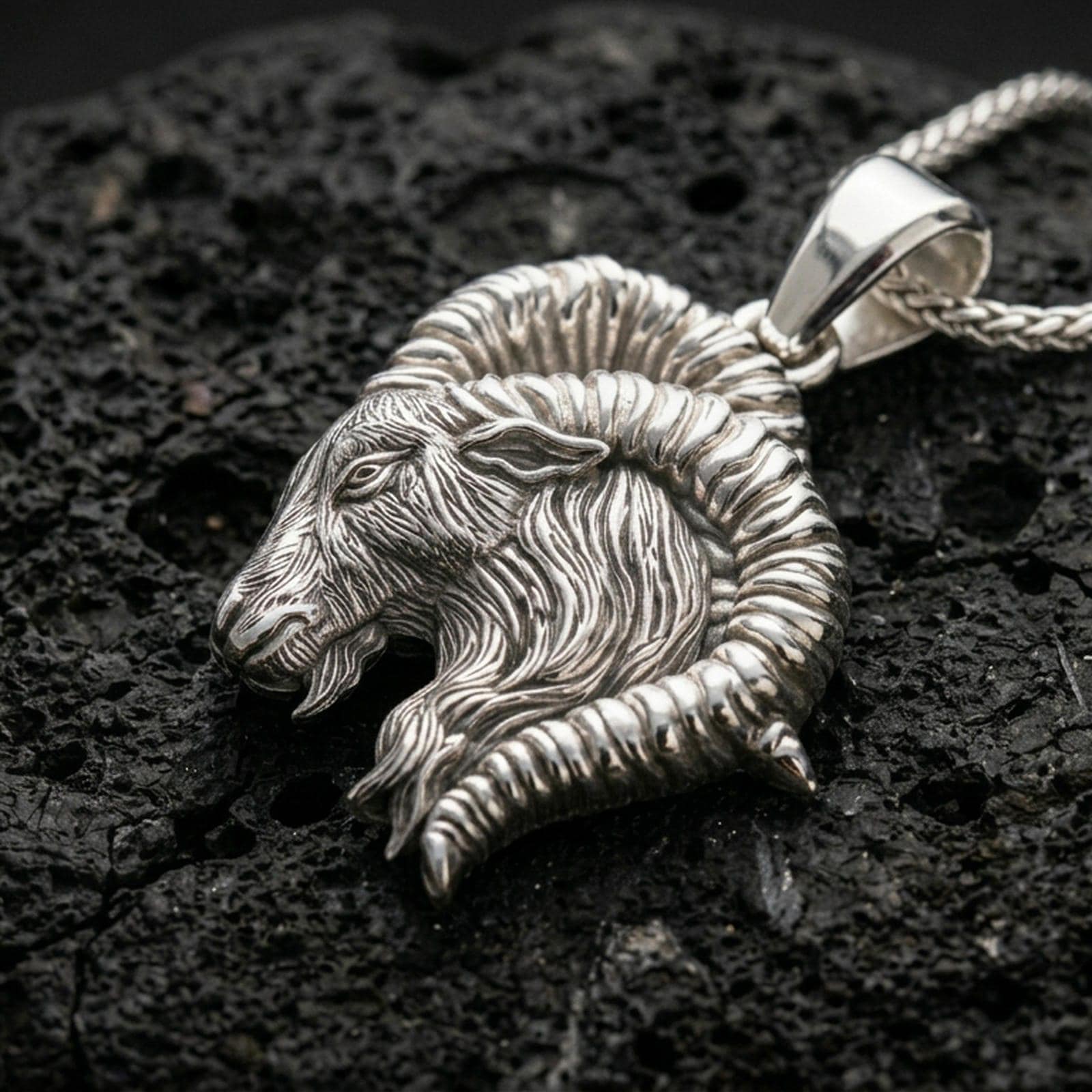 Polished silver ram necklace with sculpted Aries head, dramatic curled horns, and detailed fur texture placed on textured volcanic rock in a premium studio product shot