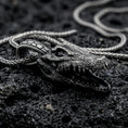 Load image into Gallery viewer, Oxidized alligator skull pendant with open jaw, detailed reptile bone texture, and fierce animal silhouette displayed on dark volcanic rock in a close-up luxury jewelry composition
