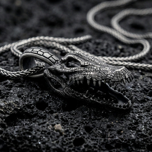 Oxidized alligator skull pendant with open jaw, detailed reptile bone texture, and fierce animal silhouette displayed on dark volcanic rock in a close-up luxury jewelry composition