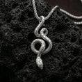Load image into Gallery viewer, Polished silver serpent necklace with elegant winding snake form, sculpted body curves, and textured scale detailing placed on textured volcanic rock in a premium studio product shot
