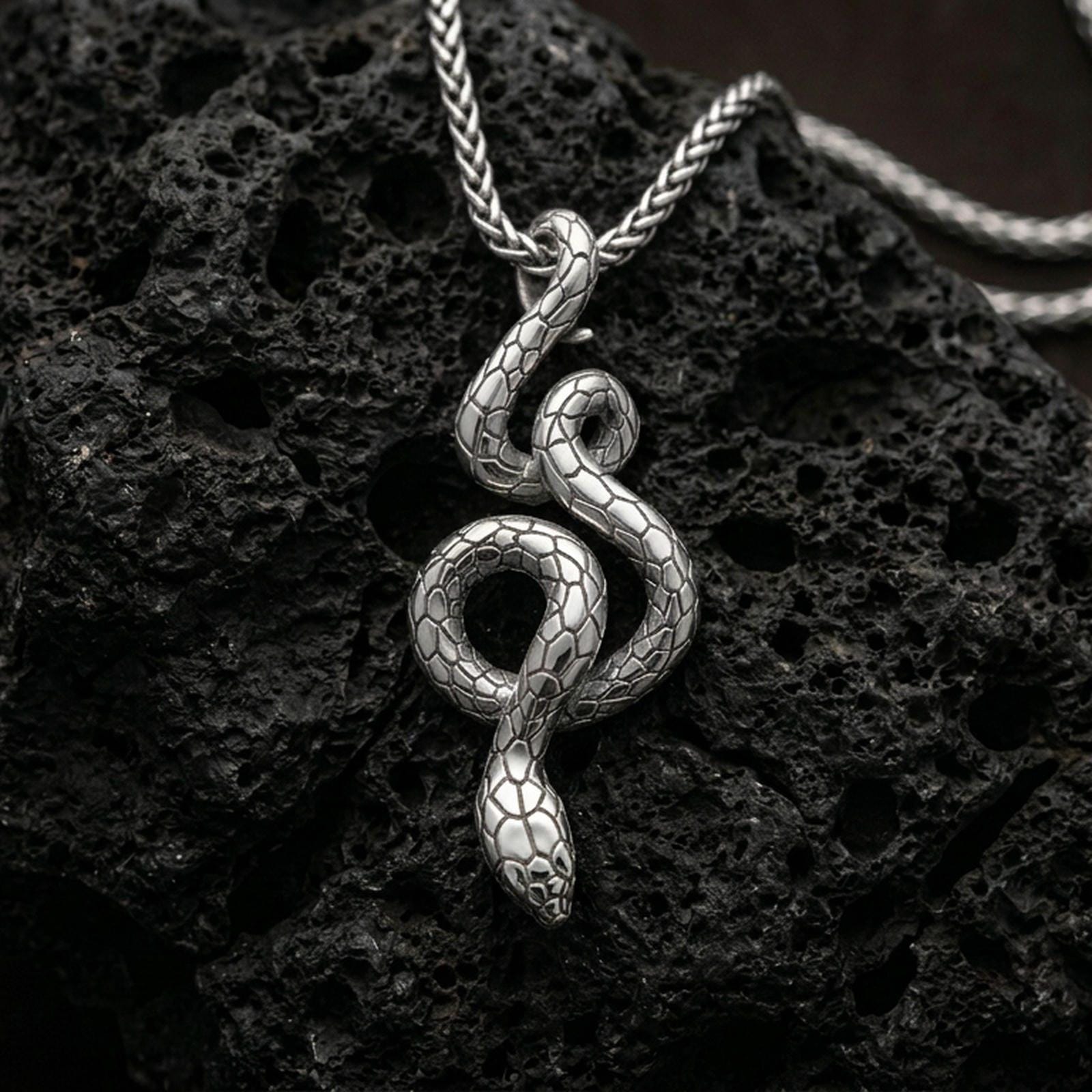 Polished silver serpent necklace with elegant winding snake form, sculpted body curves, and textured scale detailing placed on textured volcanic rock in a premium studio product shot
