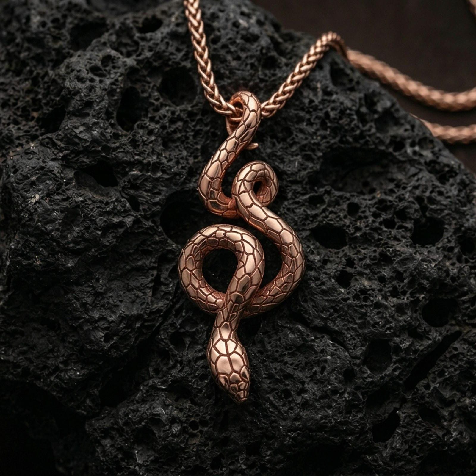 Rose gold plated serpent necklace with slender coiled snake design, detailed body texture, and elegant symbolic form resting on volcanic rock in an elegant close-up product presentation