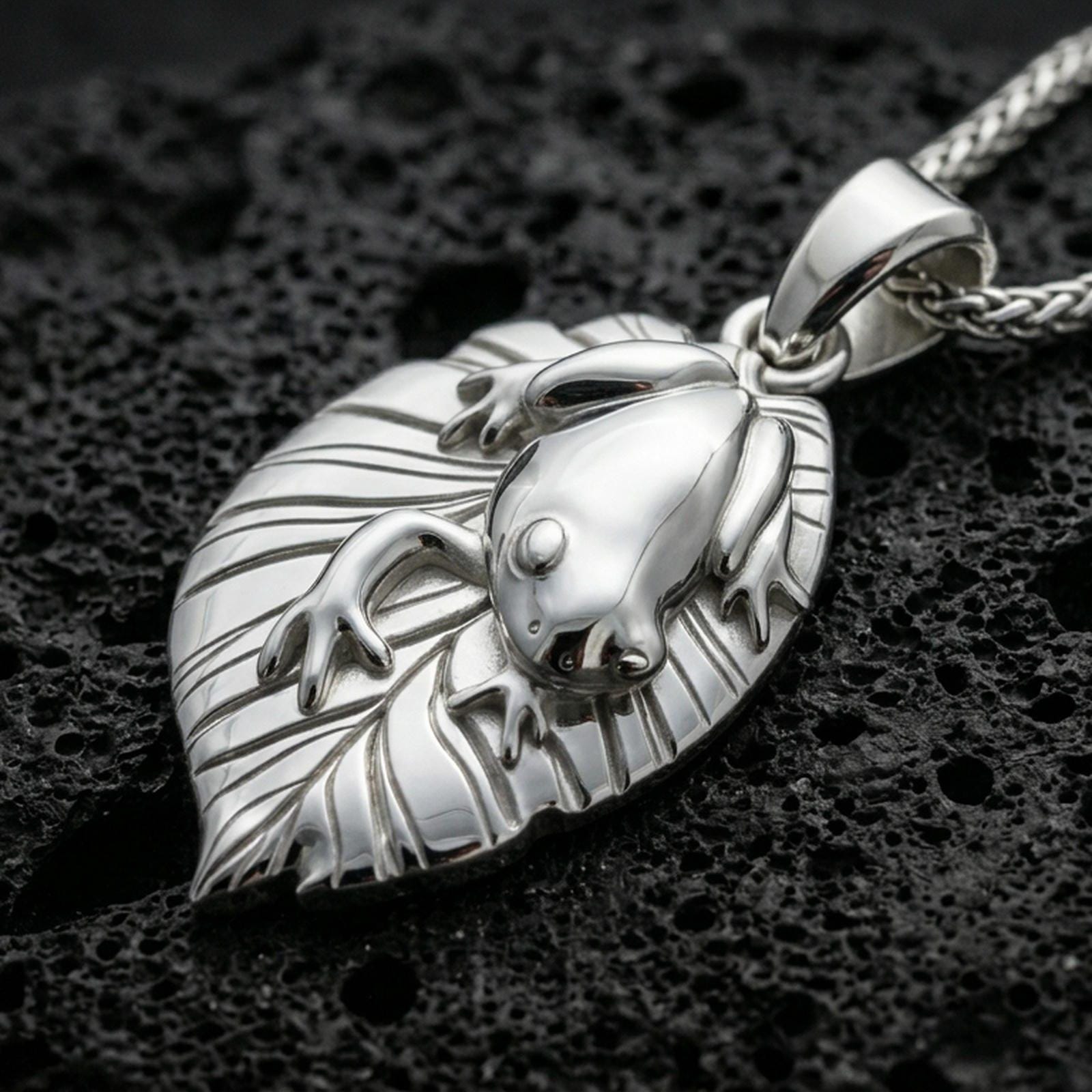 Polished silver frog necklace with sculpted frog form, botanical leaf backing, and clean organic contours placed on textured volcanic rock in a premium studio product shot