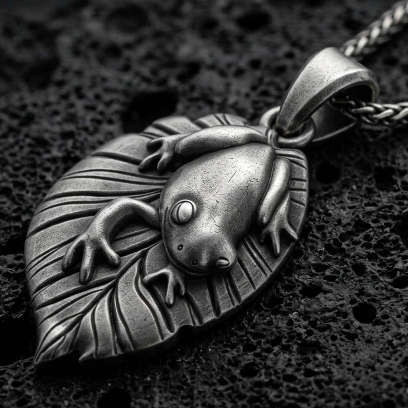 Oxidized frog pendant with smooth amphibian relief, leaf-shaped base, and nature-inspired animal silhouette displayed on dark volcanic rock in a close-up luxury jewelry composition