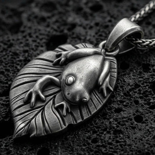 Oxidized frog pendant with smooth amphibian relief, leaf-shaped base, and nature-inspired animal silhouette displayed on dark volcanic rock in a close-up luxury jewelry composition