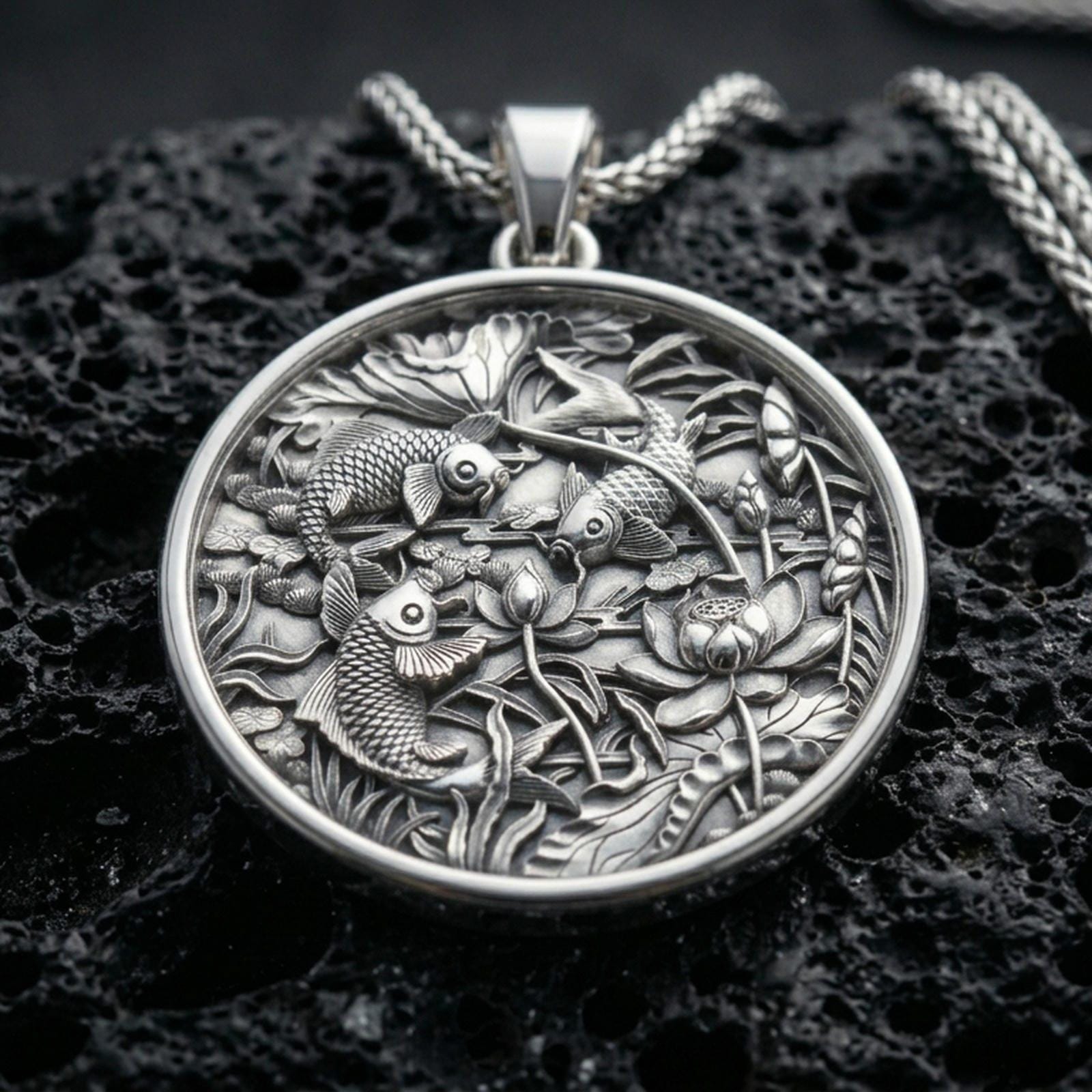 Polished silver koi fish necklace with intricate lotus flower details, layered pond elements, and circular nature-inspired design placed on textured volcanic rock in a premium studio product shot