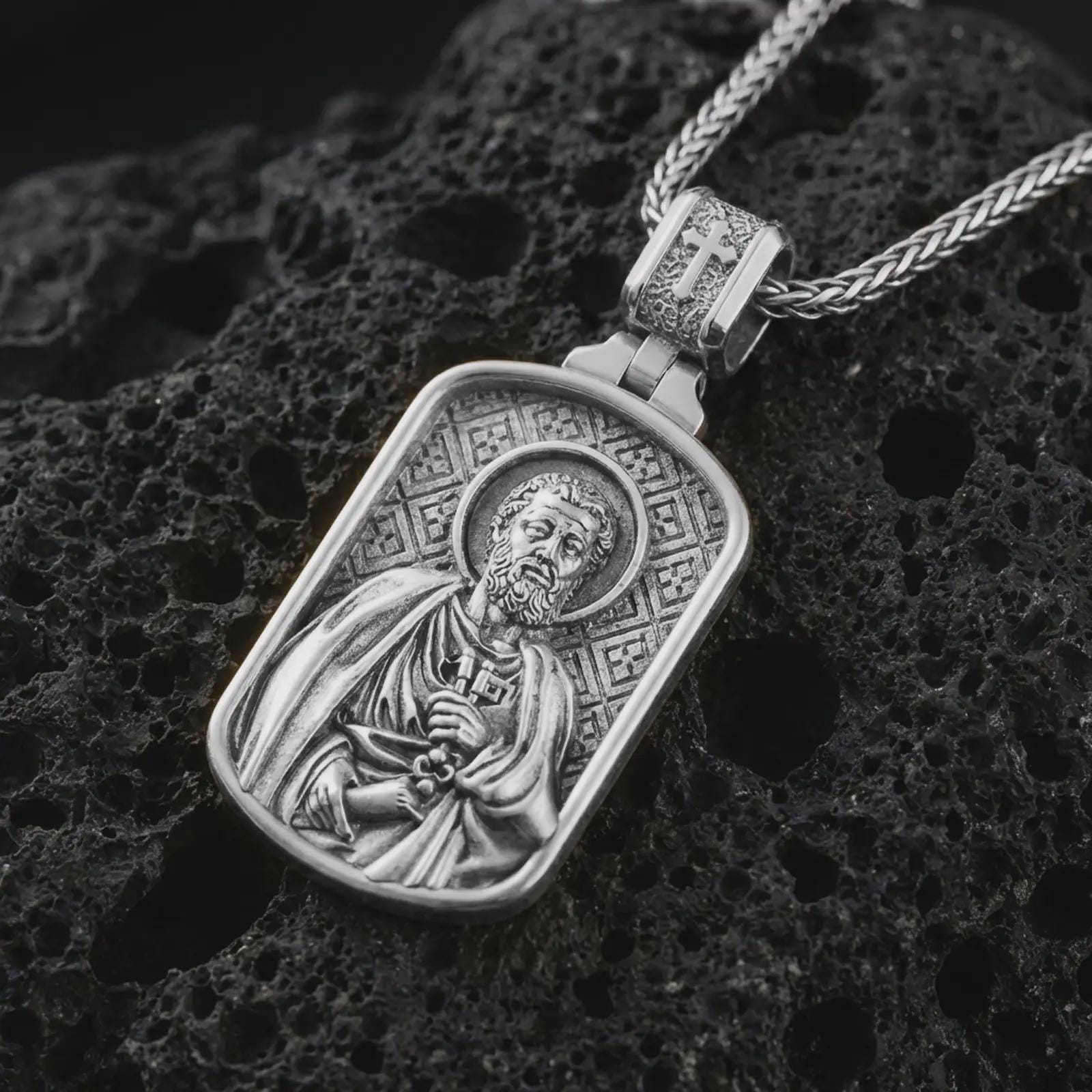 Polished silver Saint Peter dog tag pendant necklace displayed on black volcanic rock, shown in a refined macro product shot with detailed saint figure, key, and engraved background pattern.
