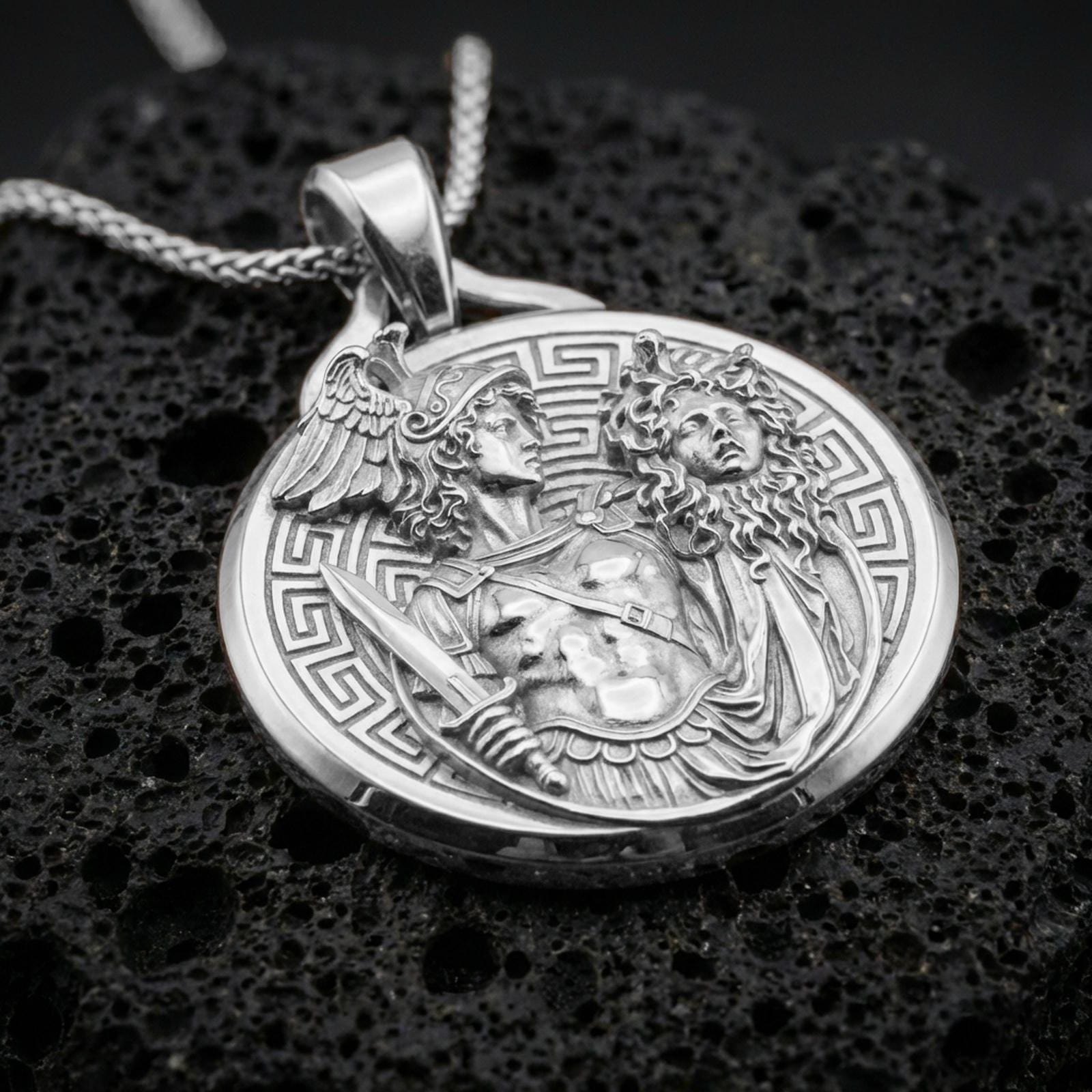 A polished 925 sterling silver Greek mythology coin pendant on volcanic rock featuring Perseus holding the Medusa head.