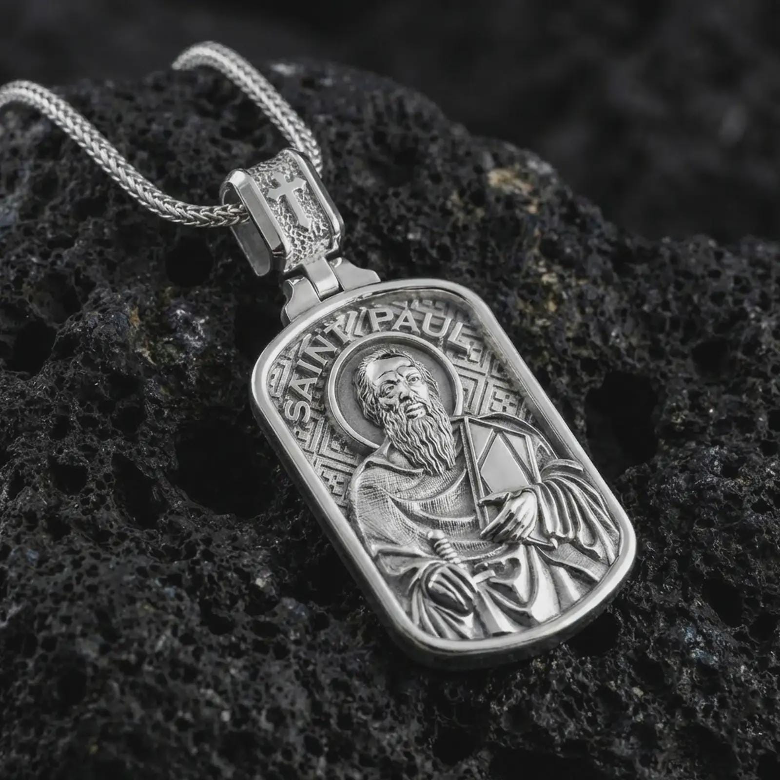 Polished silver Saint Paul dog tag pendant necklace displayed on black volcanic rock, shown in a refined macro product shot with detailed saint figure, sword, and engraved lettering.