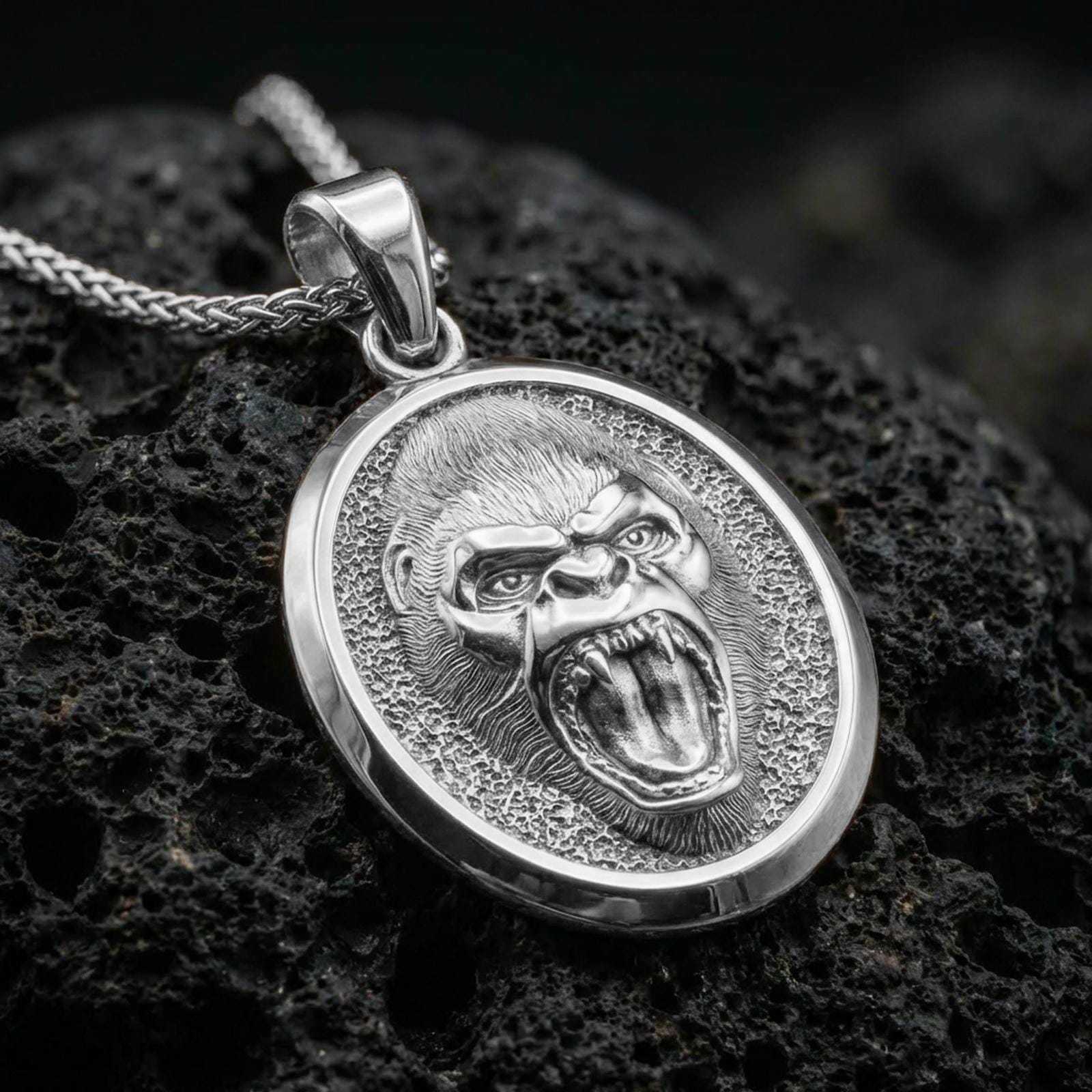 Polished silver gorilla necklace with detailed roaring primate face, sculpted fur texture, and oval statement medallion design placed on textured volcanic rock in a premium studio product shot