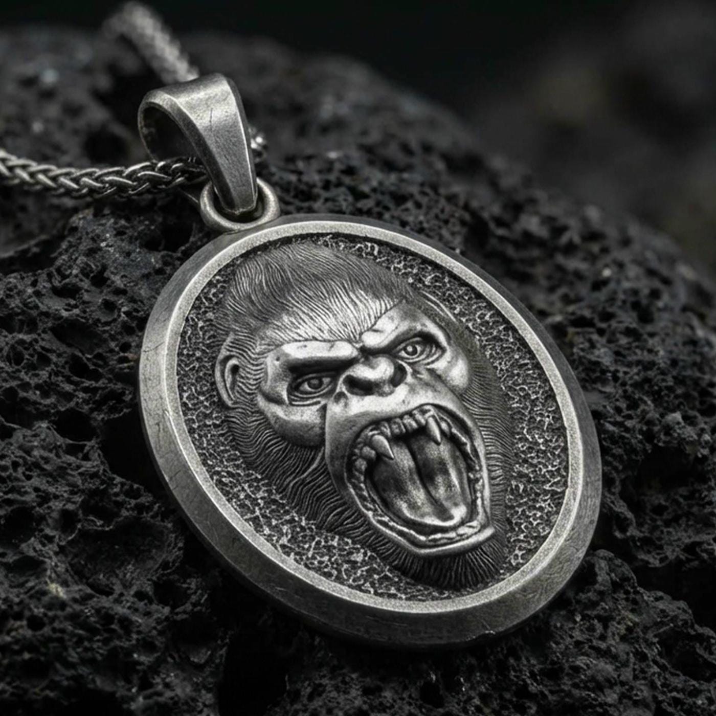 Oxidized roaring gorilla pendant with fierce open-mouth ape relief, oval medallion frame, and bold primal animal silhouette displayed on dark volcanic rock in a close-up luxury jewelry composition