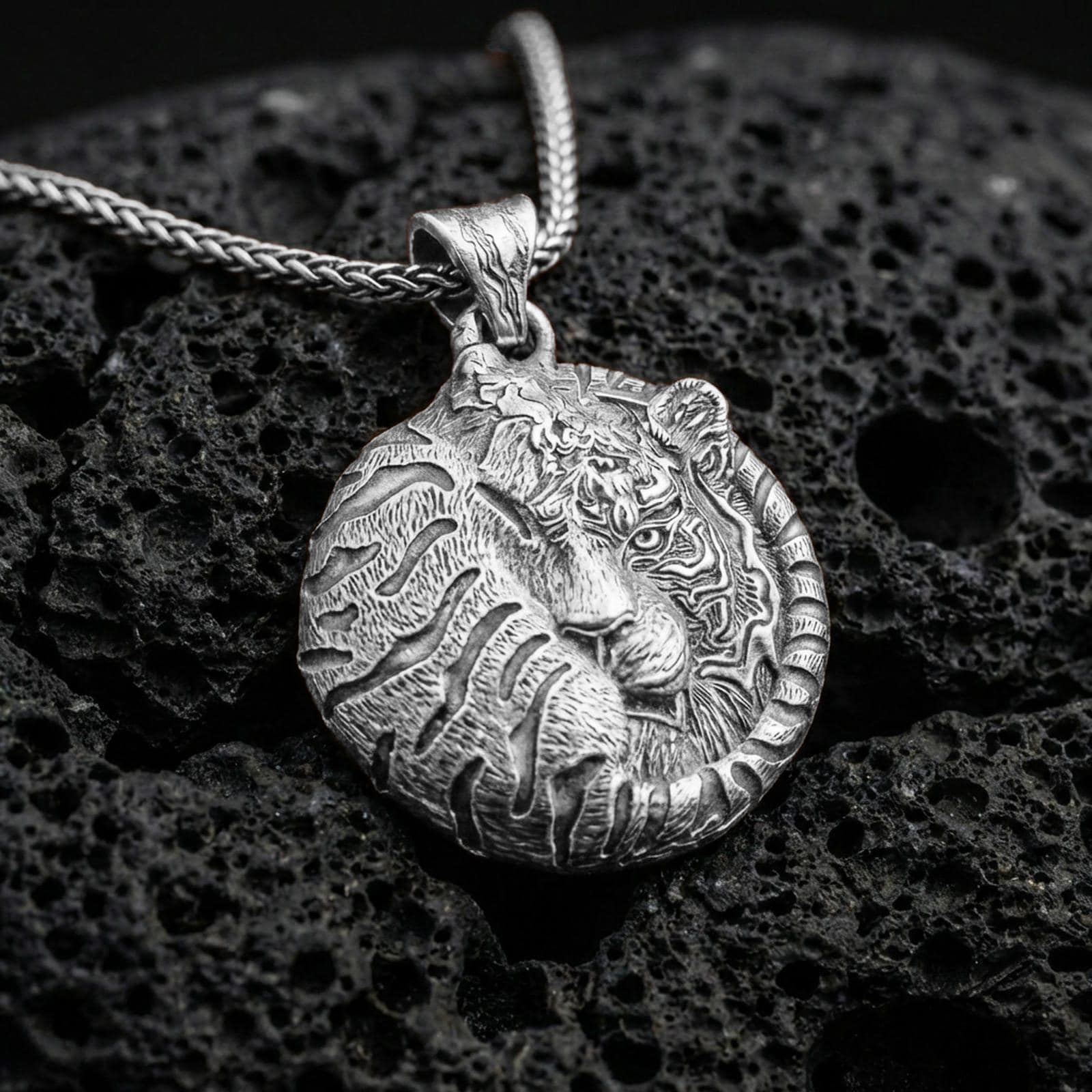 Polished silver tiger necklace with resting wild cat design, intricate facial detailing, and wrapped tail composition placed on textured volcanic rock in a premium studio product shot