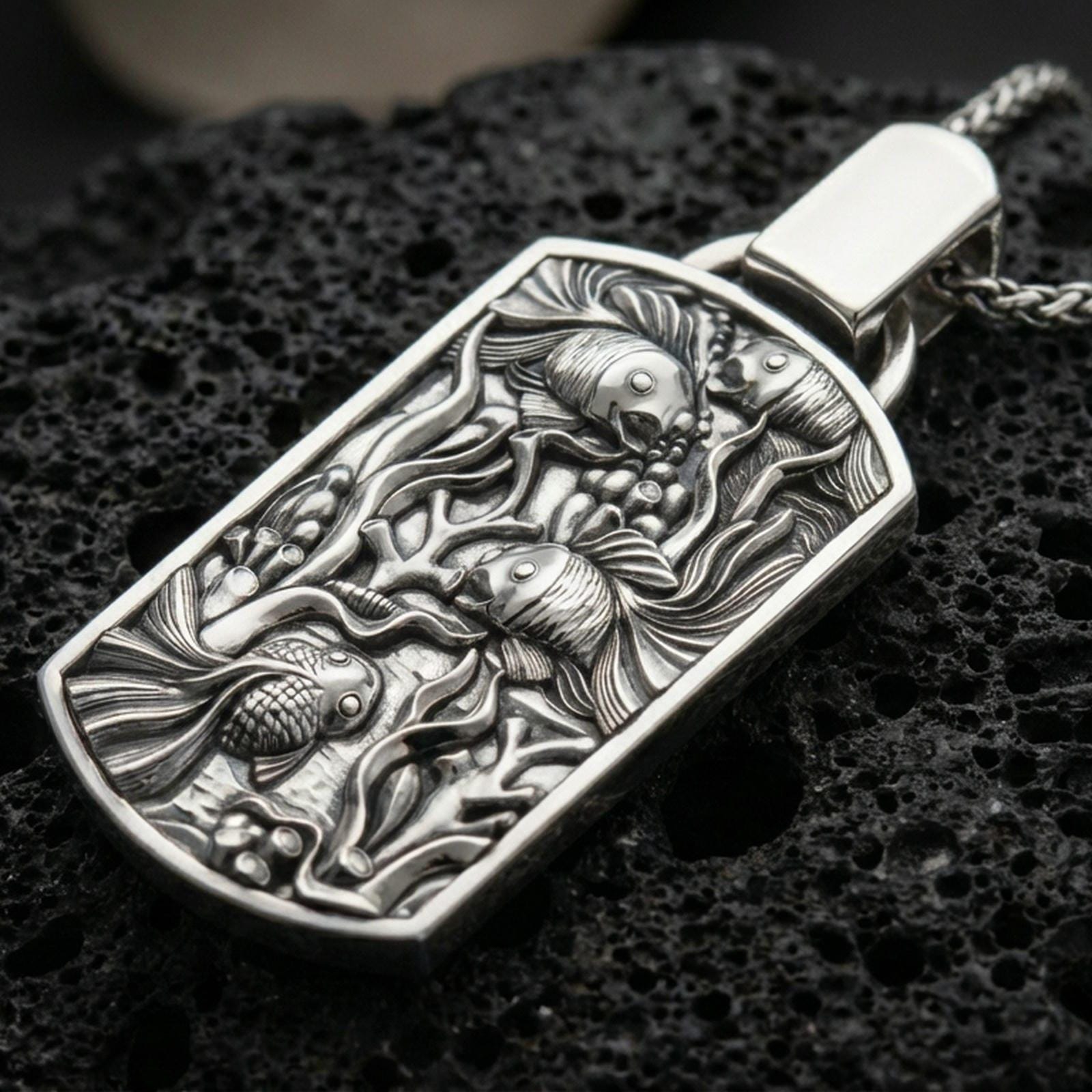 Polished silver aquarium necklace with sculpted fish details, flowing underwater plant motifs, and refined rectangular medallion design placed on textured volcanic rock in a premium studio product shot