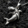 Load image into Gallery viewer, Oxidized shark pendant with open-mouth predator design, sculpted body curve, and playful marine silhouette displayed on dark volcanic rock in a close-up luxury jewelry composition
