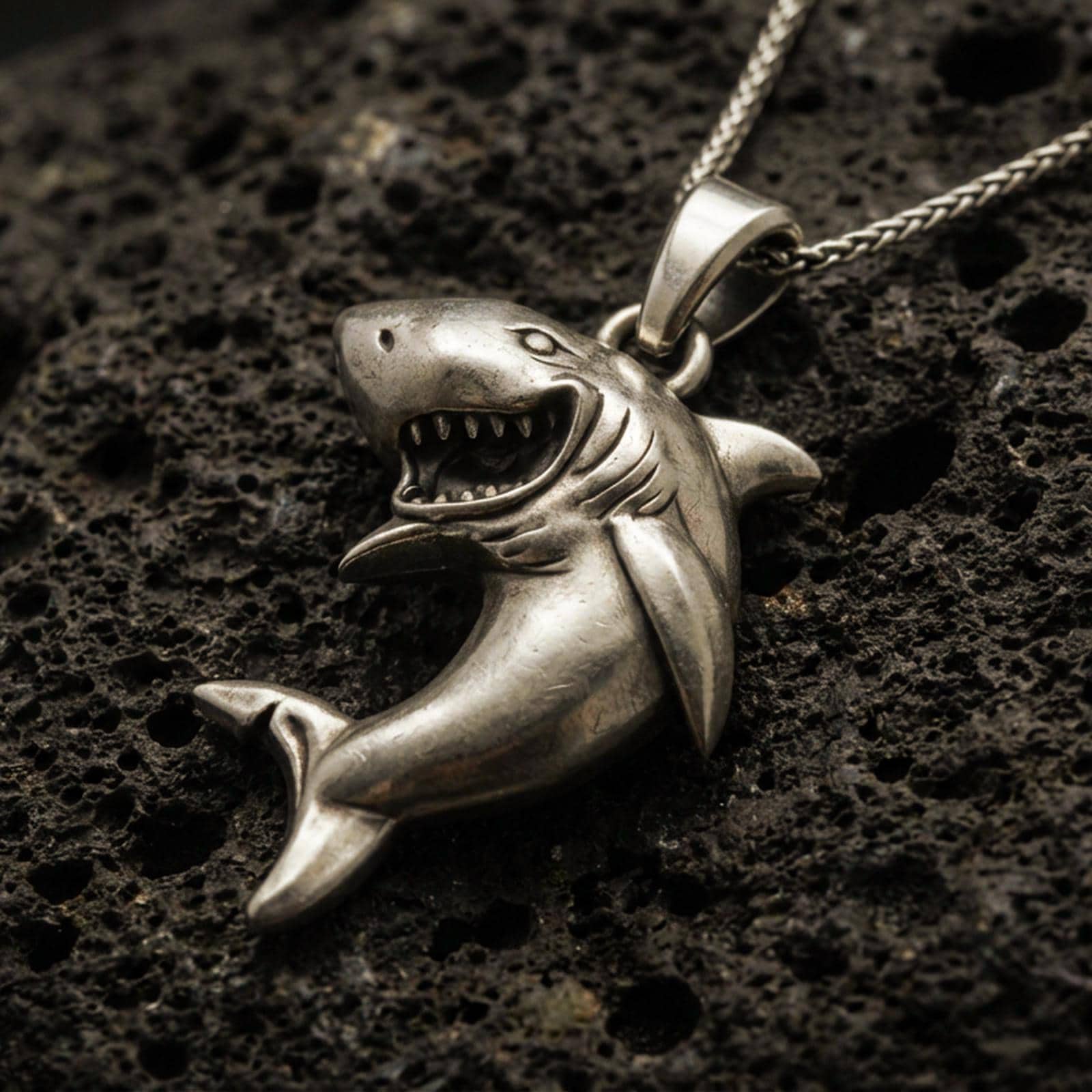 Bronze shark pendant with open-mouth predator design, sculpted body curve, and playful marine silhouette displayed on dark volcanic rock in a close-up luxury jewelry composition