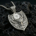Load image into Gallery viewer, Polished silver stag necklace with Celtic knot detailing, shield-inspired frame, and engraved Nordic symbol placed on textured volcanic rock in a premium studio product shot

